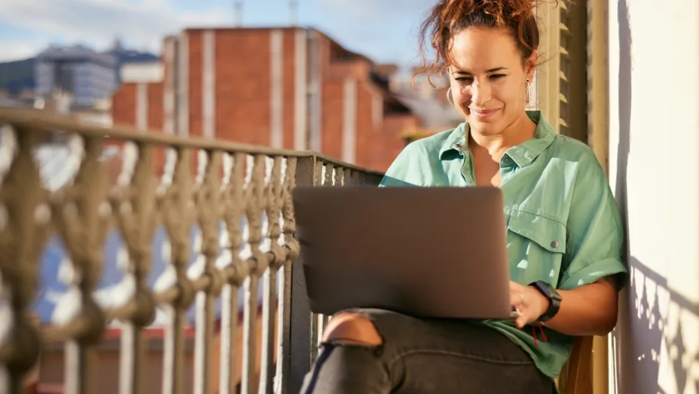 Young woman sitting on balcony using laptop with city buildings in background