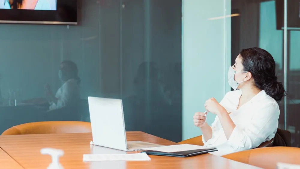 Woman sitting at conference table with laptop, participating in video call on screen