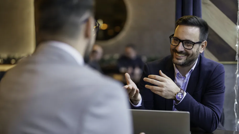Two men in stylish suits sit at a table discussing how to manage a team with a laptop on the table between them.