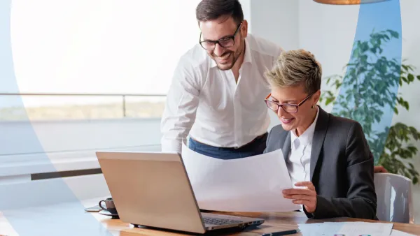 Two business professionals collaborating at a desk, reviewing documents and working together on a laptop.