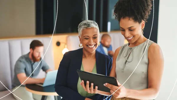 Two businesswomen standing together and smiling while looking at a tablet, with colleagues working in the background.
