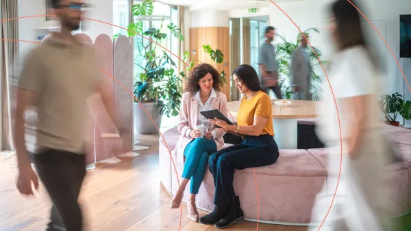 Two professionals seated on a bench in a modern open office, discussing information on a tablet while others walk by.
