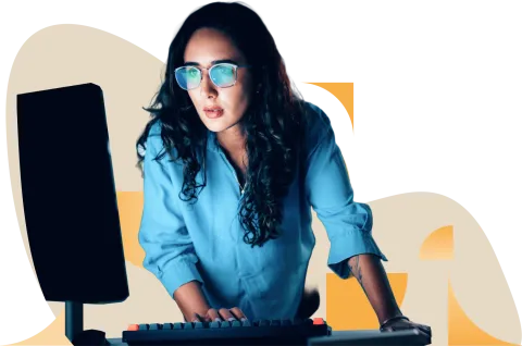 Woman with long dark hair wearing blue shirt and glasses, sitting at desk with computer monitor