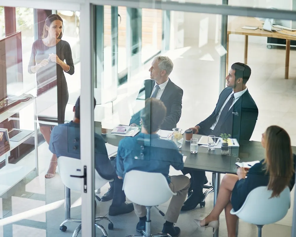 People seated at desks in modern glass-walled office space having a business meeting