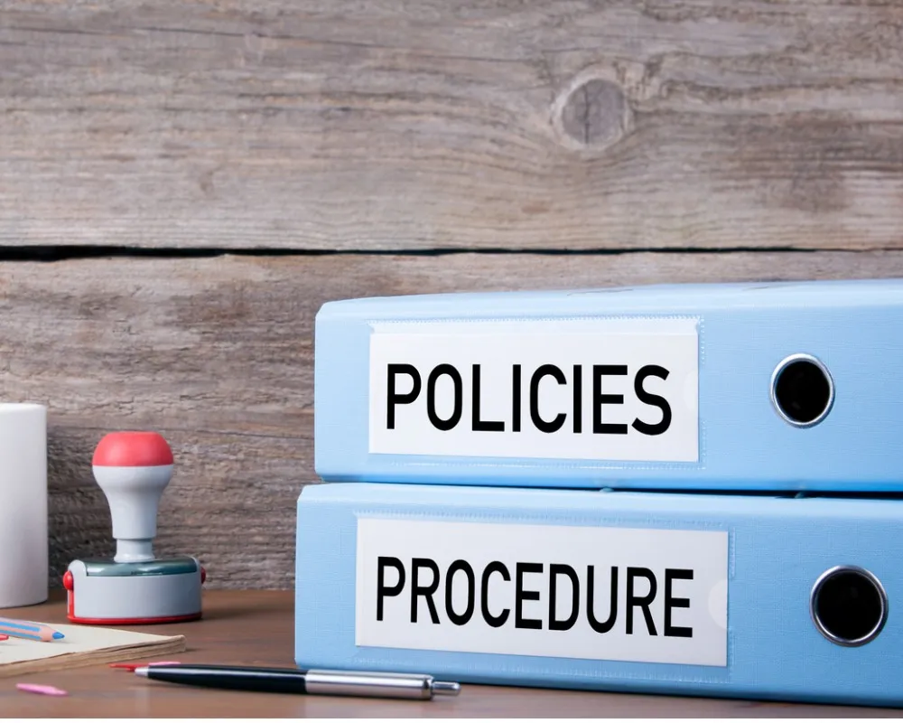 Two blue binders labeled 'Policies' and 'Procedure' stacked on wooden desk with office supplies