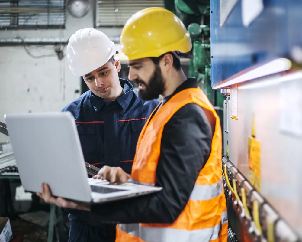 Two construction workers in safety gear reviewing plans on a laptop outdoors