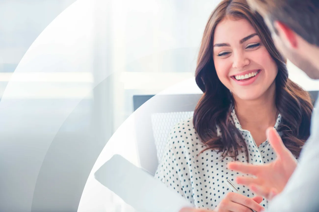 A woman smiling while talking with a colleague, holding a pen and document