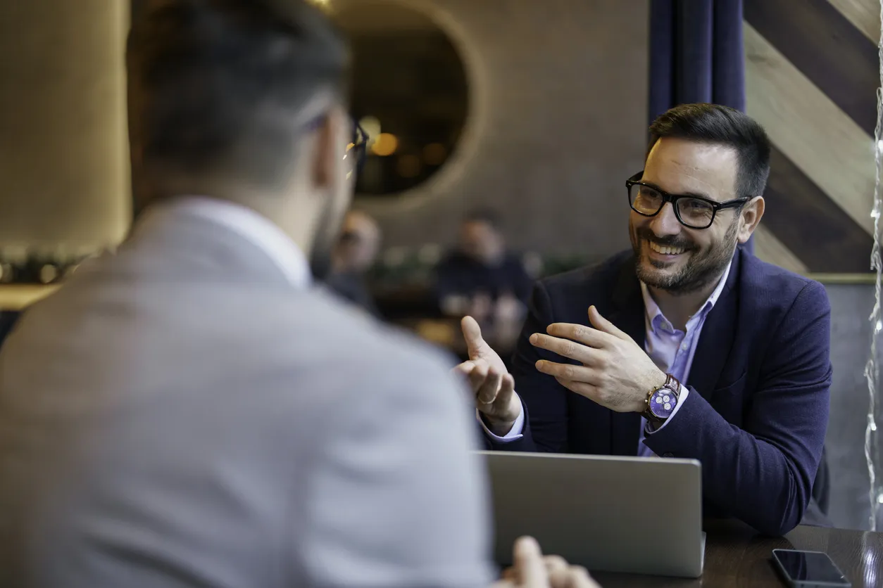 Two men in stylish suits sit at a table discussing how to manage a team with a laptop on the table between them.