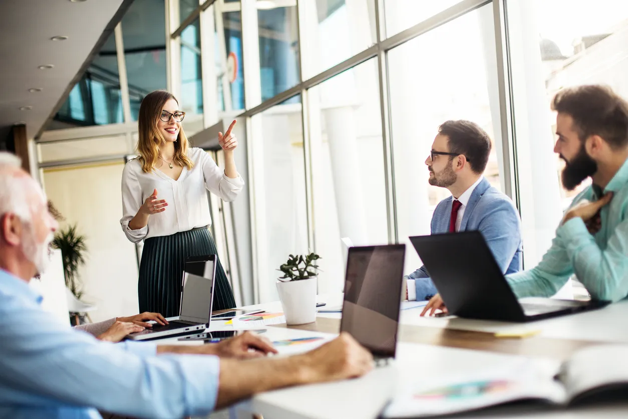 Men sat at a meeting room table with open laptops focus their attention on a woman who stands as she explains coaching as a manager.