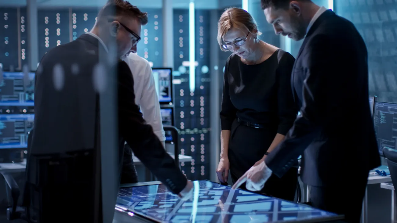 Three IT Service Managers in business wear look down at a large screen in a blue lit room