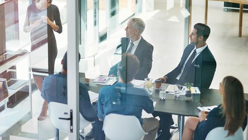 People seated at desks in modern glass-walled office space having a business meeting
