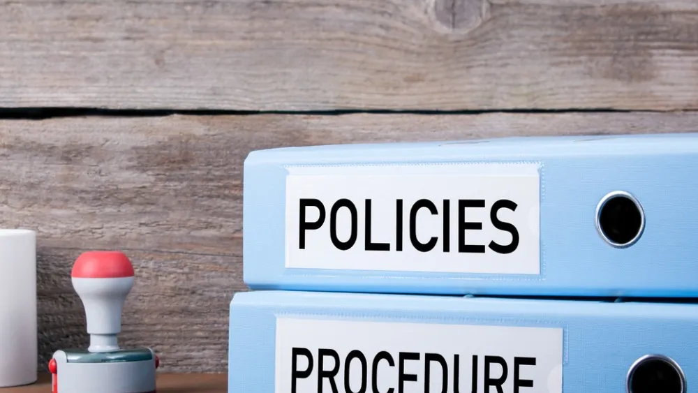 Two blue binders labeled 'Policies' and 'Procedure' stacked on wooden desk with office supplies