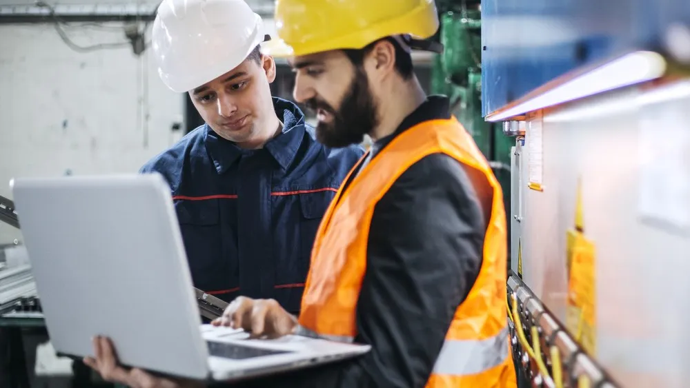 Two construction workers in safety gear reviewing plans on a laptop outdoors