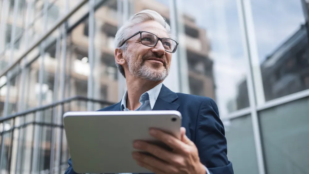 Smiling businessman in glasses using tablet device outside modern office building