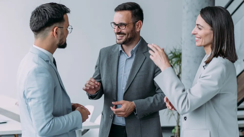 Three professionals in suits smiling and talking in a modern office