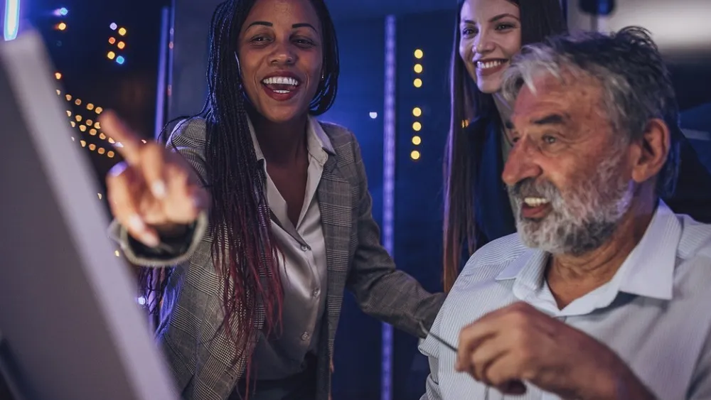 Three people smiling and interacting around a computer in a dimly lit room
