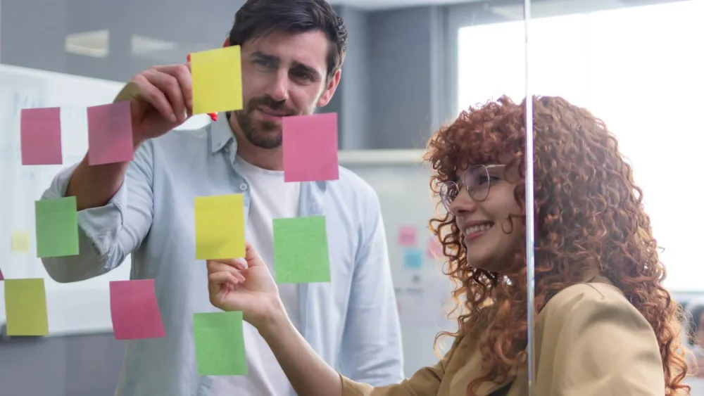 Two colleagues discussing colorful sticky notes on a glass wall in a modern office