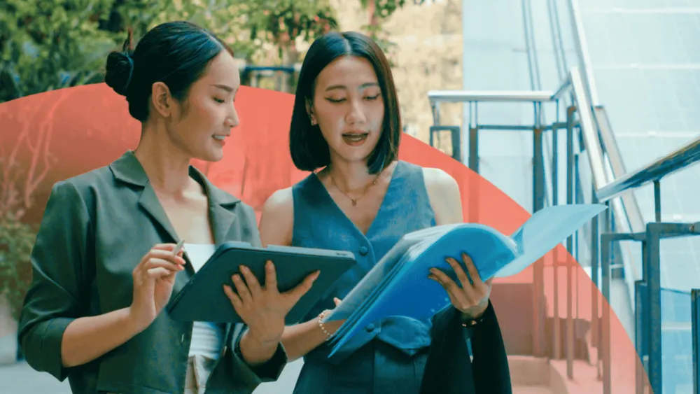 Two female professionals stand outside at the bottom of a set of stairs. One is holding a tablet and the other is holding a folder. They look to be discussing the contents of the folder.