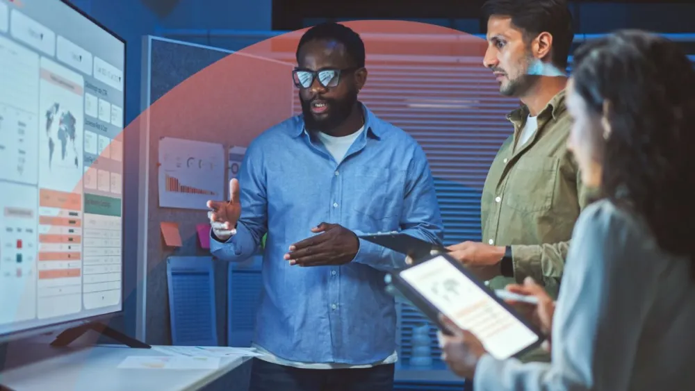 Man in blue shirt presenting to two colleagues in modern office with digital displays
