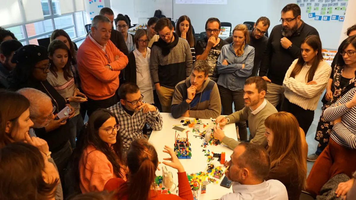 A crowded room, people surround a table with lego pieces on it