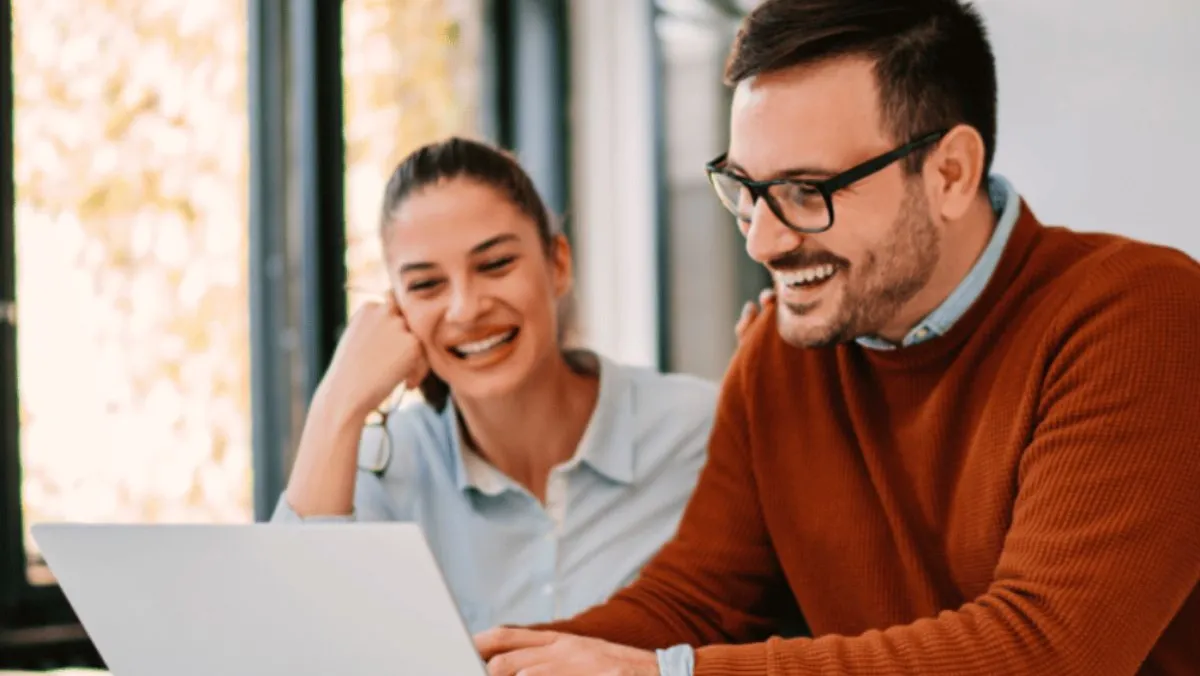 Two people smiling while looking at a laptop together in an office setting