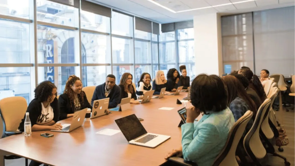Team of professionals sitting around a wooden conference table during a meeting in a bright, modern office with large windows.