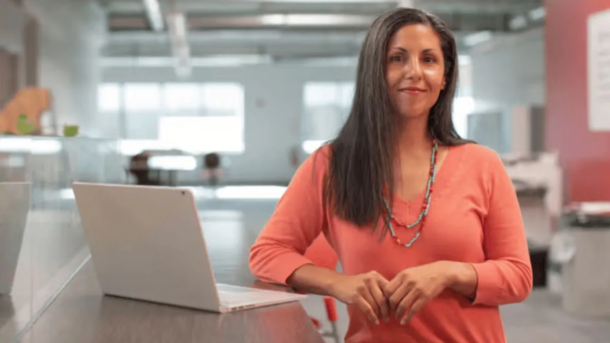 Woman in coral blouse standing by a desk with laptop in modern office setting.