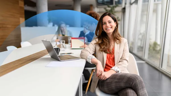 Confident businesswoman sitting at a meeting table, smiling at the camera, with colleagues in the background.