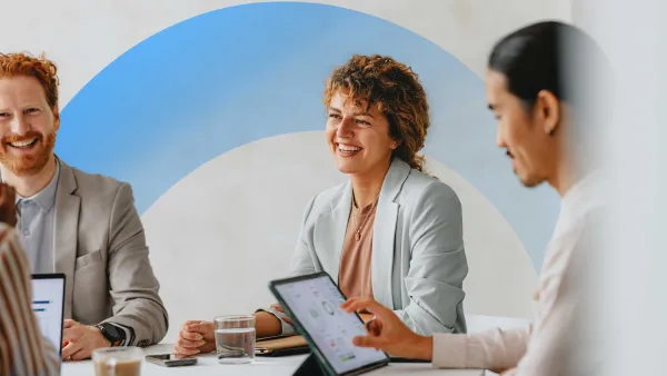 Team members smiling and collaborating during a meeting, using digital tablets and laptops in a bright office.