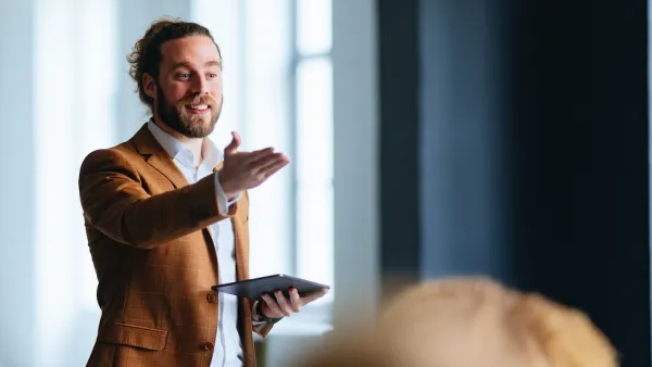 Man in brown jacket speaking and gesturing during a presentation