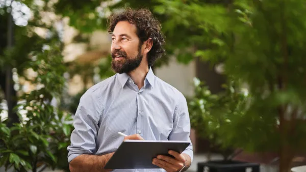 Man wearing a light blue shirt, holding a clipboard, looks to his right. In the background are green trees and plants in soft focus.